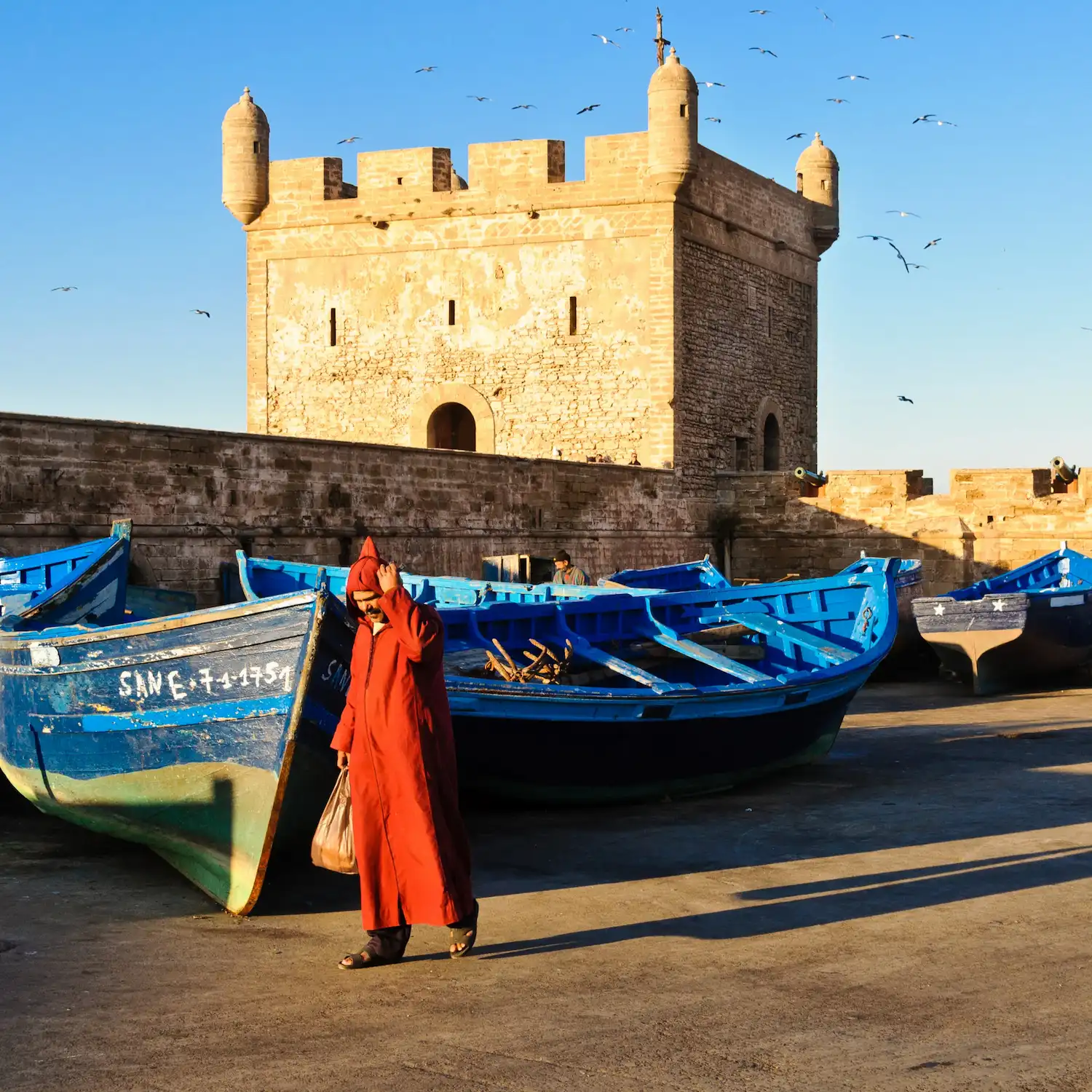 Blue fishing boats near historic fortress of Essaouira in Morocco with a Man in a red Jellaba during a tour with Moroccan Guides Travel
