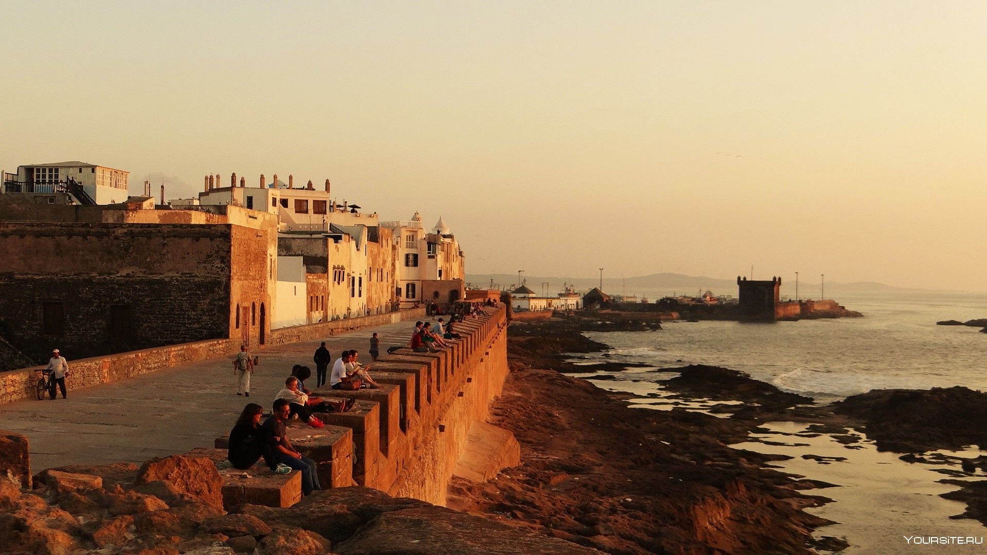 Sunset over coastal Essaouira fortress wall with people sitting along rocky shoreline