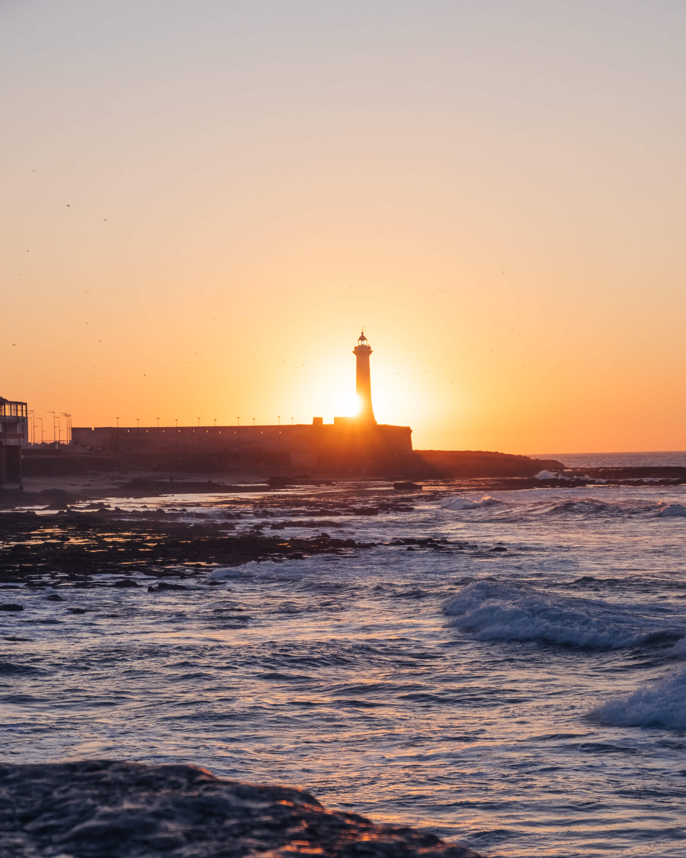 Lighthouse in Rabat Morocco silhouetted against golden sunset with rocky ocean waves during a guided tour Moroccan Guides travel
