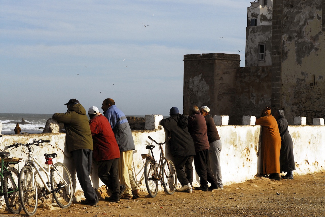 People with bicycles looking out over stormy sea near old stone wall oof Essaouira in Morocco