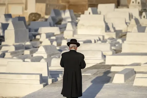 Rear view of a Jewish man wearing a traditional black hat and coat walking through a historic white-tombstone Jewish cemetery in Morocco with Moroccan Guides Travel.