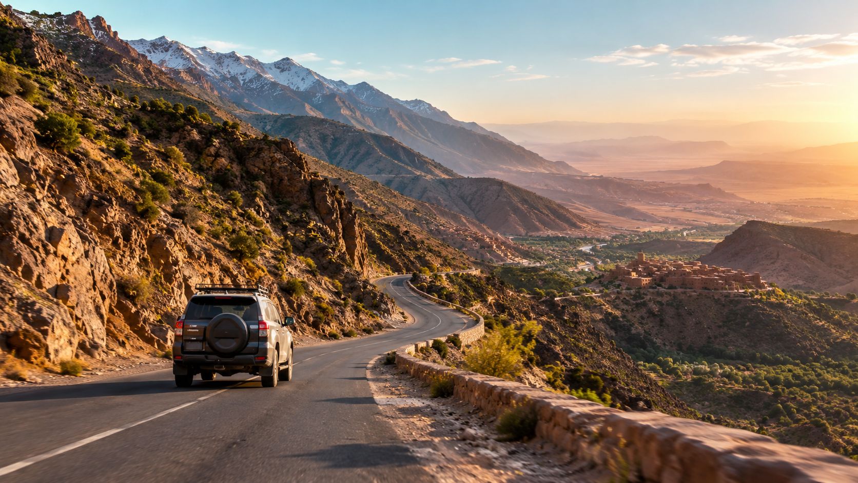 A gray SUV driving on a winding mountain road near a traditional village in Morocco at sunset.