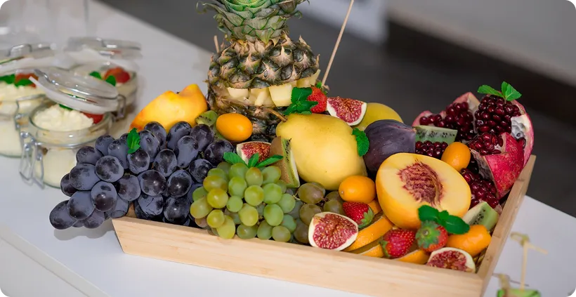 Wooden tray with assorted fresh fruits including pineapple, grapes, peach, pomegranate, figs, and strawberries.