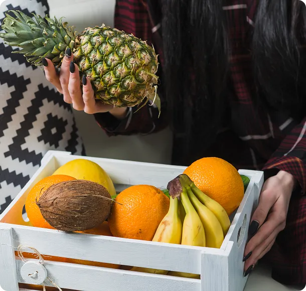 Person holding a pineapple above a white crate filled with bananas, oranges, coconut, and other fruits.