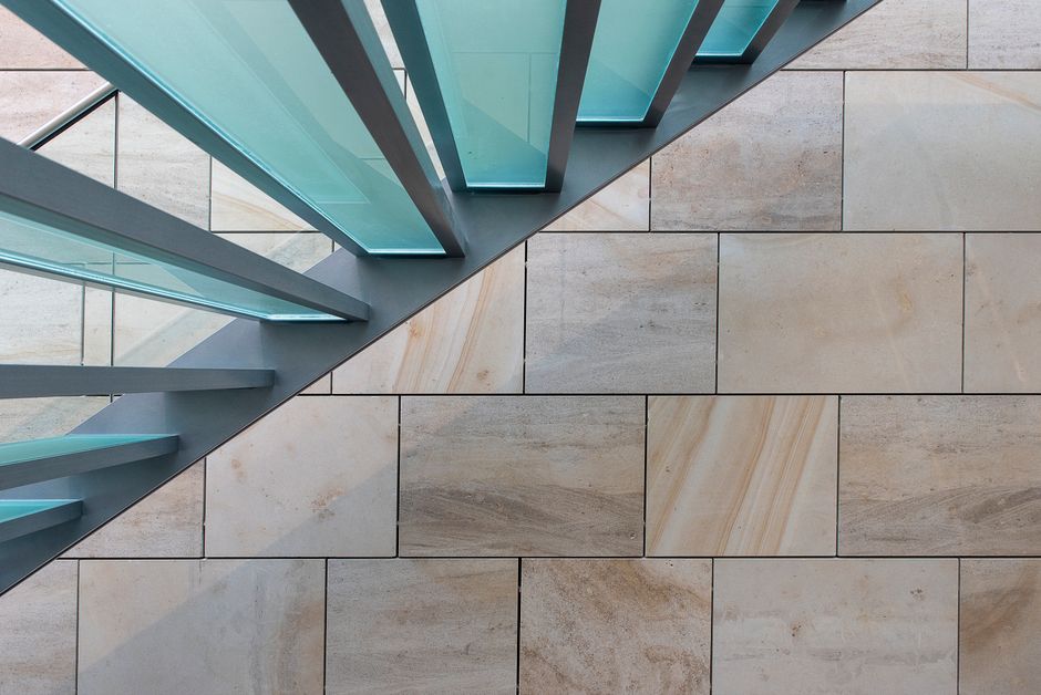 Top-down view of a modern glass and metal staircase over a tiled floor, featuring a sharp geometric design.