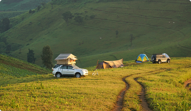 A campsite on rolling grassy hills. A car path leads you to a silver SUV with a ten on top and three regular tents. Another SUV parked in front of the last tent.