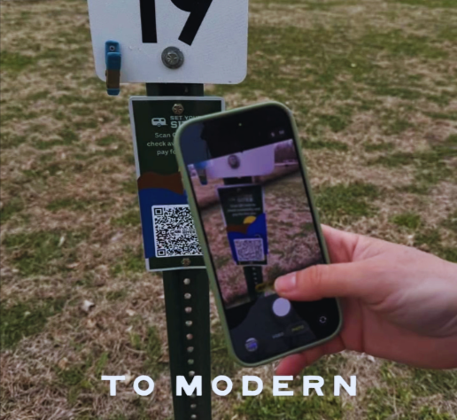 Hand holding smartphone scanning QR code on a campsite signpost in a grassy area.