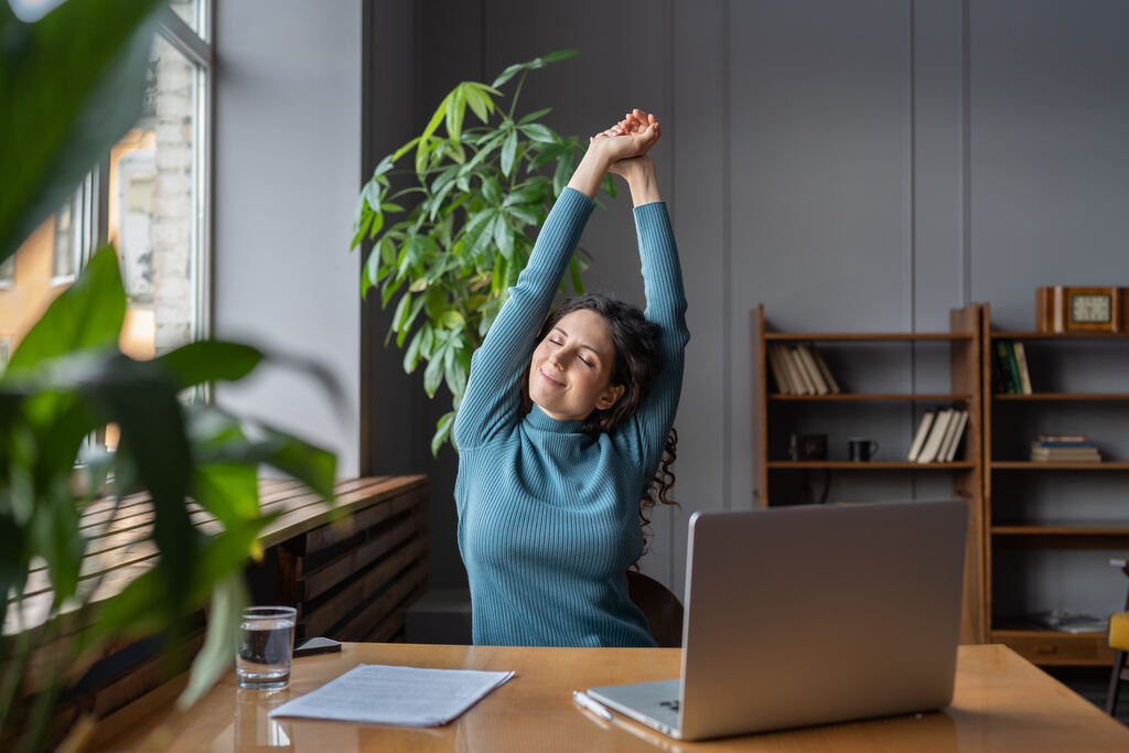 Uma mulher jovem, sentada em frente a um laptop em um ambiente de escritório, alonga os braços para cima enquanto fecha os olhos e sorri levemente, parecendo relaxada. Ela usa um suéter azul e está cercada por plantas e estantes com livros, criando uma atmosfera confortável e tranquila.