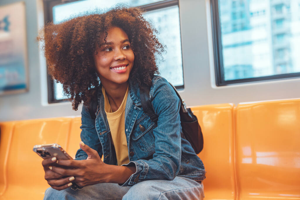 Mulher sorridente com cabelo cacheado volumoso, sentada em um assento laranja de transporte público, segurando um celular. Ela veste uma jaqueta jeans sobre uma camiseta amarela e carrega uma mochila preta nas costas. A expressão dela é relaxada e alegre, olhando para o lado com um olhar descontraído. Ao fundo, há janelas que mostram prédios embaçados.