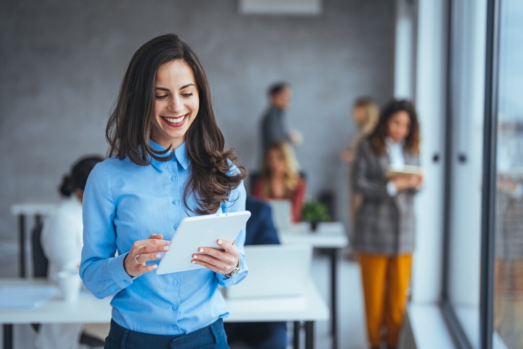 "Mulher sorridente em ambiente de escritório, usando uma camisa azul e segurando um tablet, aparentando estar concentrada e satisfeita com o que vê na tela. Ela está em primeiro plano, enquanto, ao fundo, outros colegas de trabalho conversam e trabalham em um ambiente aberto e iluminado por luz natural que entra através de grandes janelas de vidro. A atmosfera é profissional e amigável."