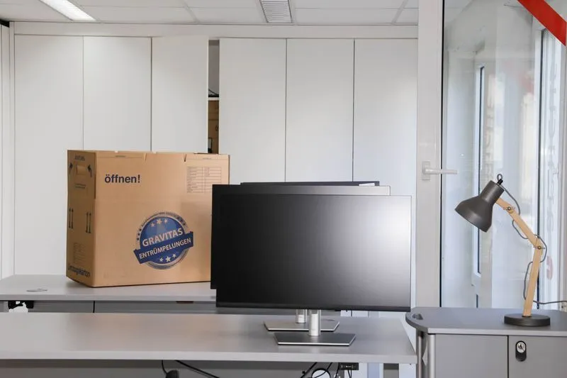Ein leergeräumter Büroraum mit Schreibtischen und Bildschirmen, daneben ein beschrifteter GRAVITAS-Umzugskarton.