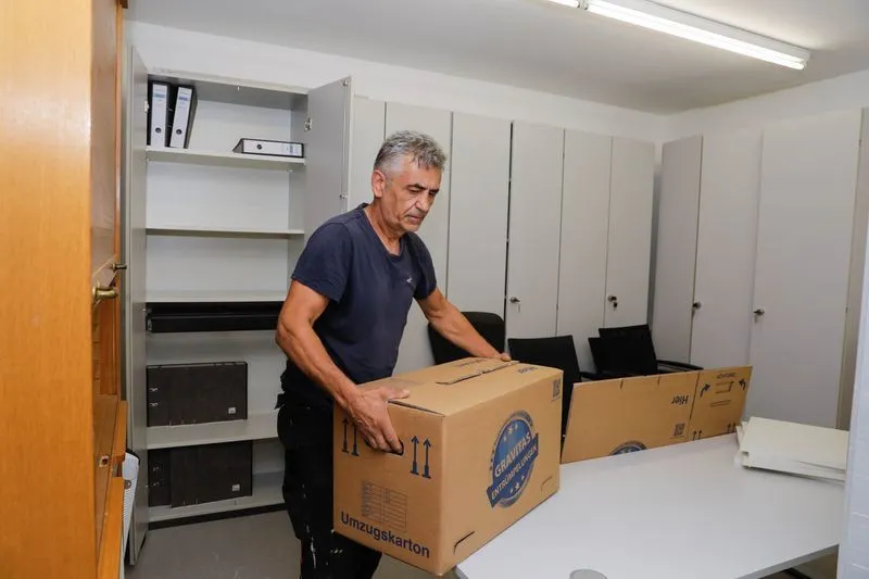 Ein Mitarbeiter trägt einen Umzugskarton mit dem Logo der Entrümpelungsfirma GRAVITAS durch ein fast leergeräumtes Büro mit geöffneten Aktenschränken.