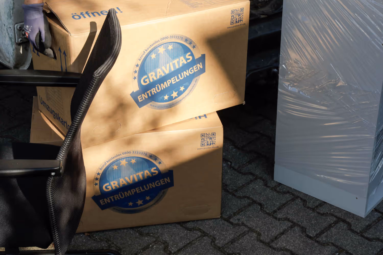 Gestapelte Umzugskartons mit GRAVITAS-Logo stehen neben einem Bürostuhl im Freien, bereit für den Abtransport.