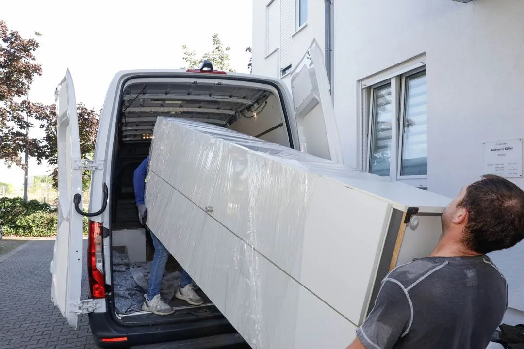 Zwei Mitarbeiter tragen ein mit Schutzfolie verpacktes Möbelstück aus einem Gebäude und verladen es in einen Transporter.
