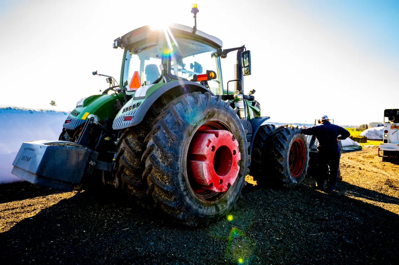 Mechanic performing tractor tire repair on large green machine with red rims under bright sunlight.