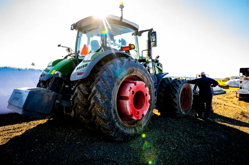 Mechanic performing tractor tire repair on large green machine with red rims under bright sunlight.