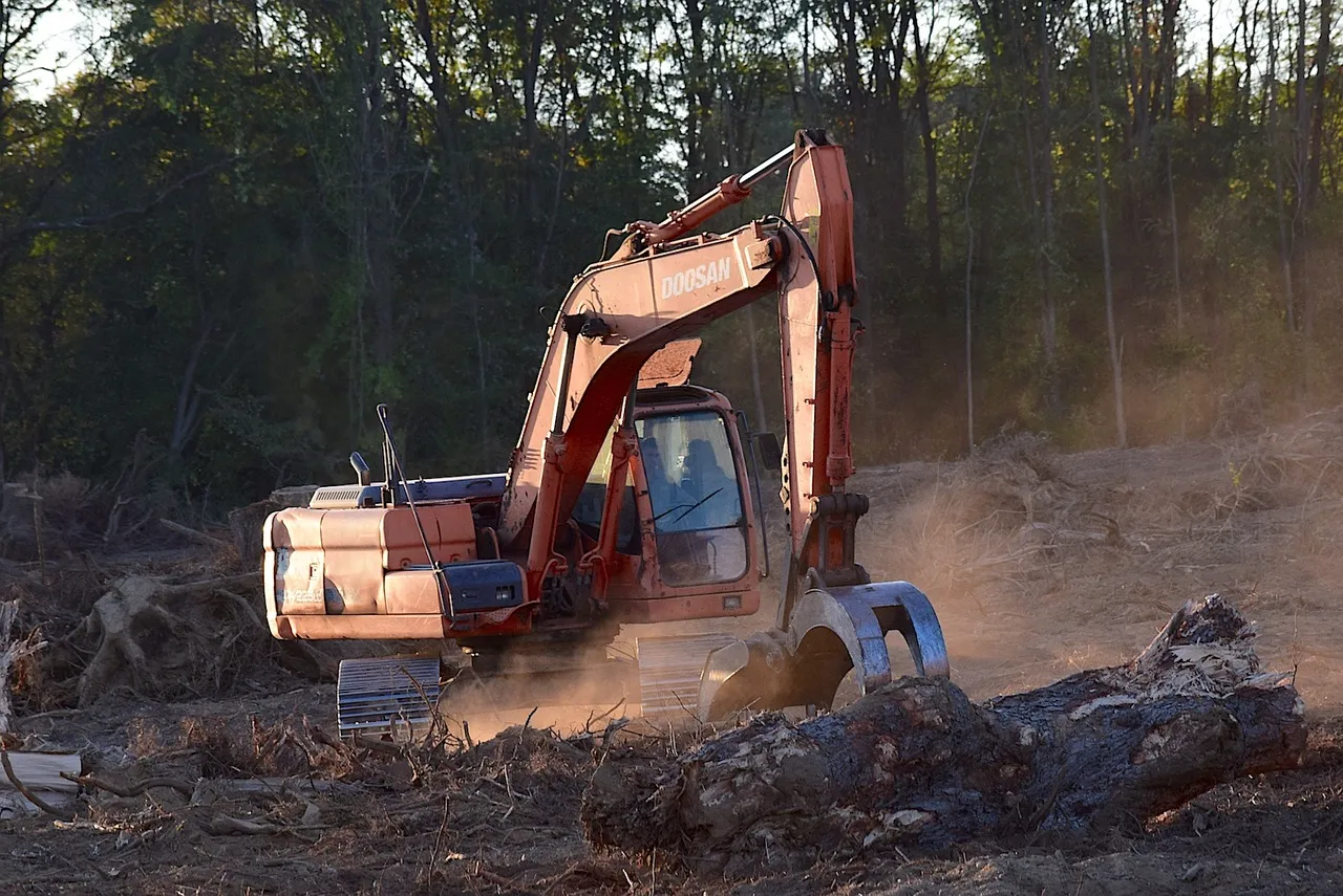Doosan excavator with grapple attachment clearing tree debris on a dusty construction site near the woods.