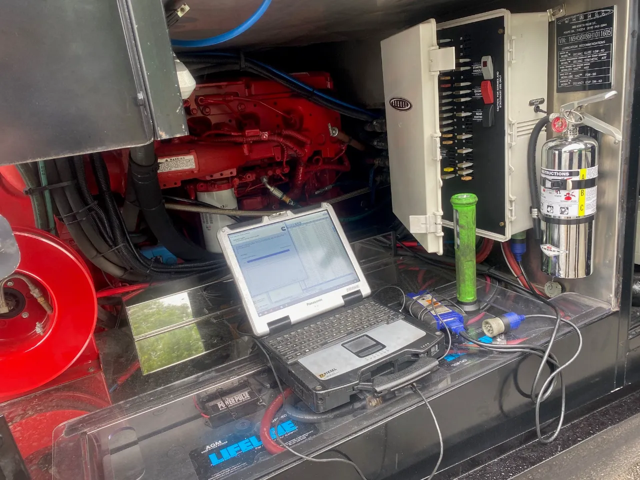 Engine diagnostics service with laptop connected to red diesel engine bay beside breaker panel and “INSTRUCTIONS” label.