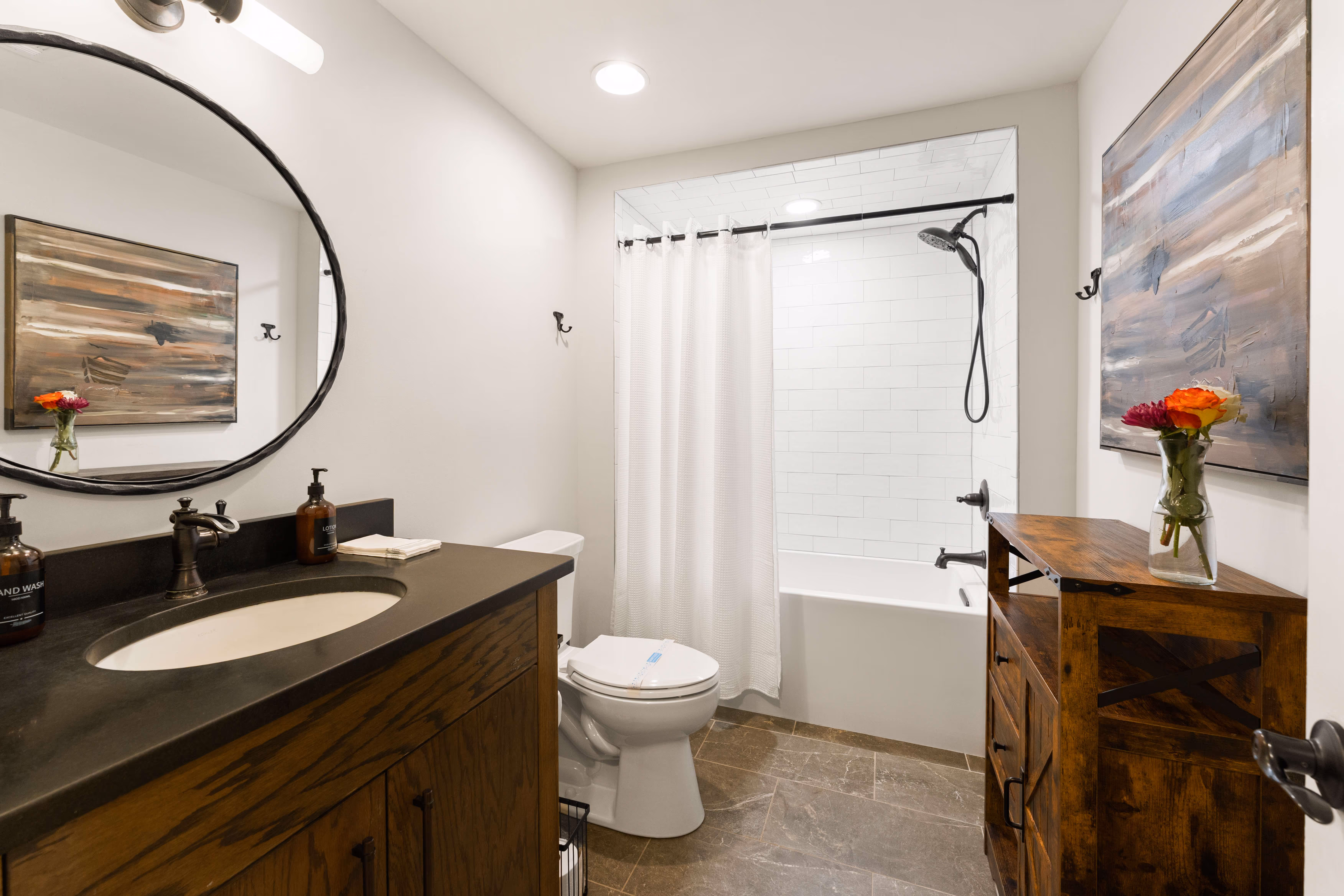 Modern bathroom with wooden vanity, round mirror, white shower curtain, and a wooden cabinet with flowers.