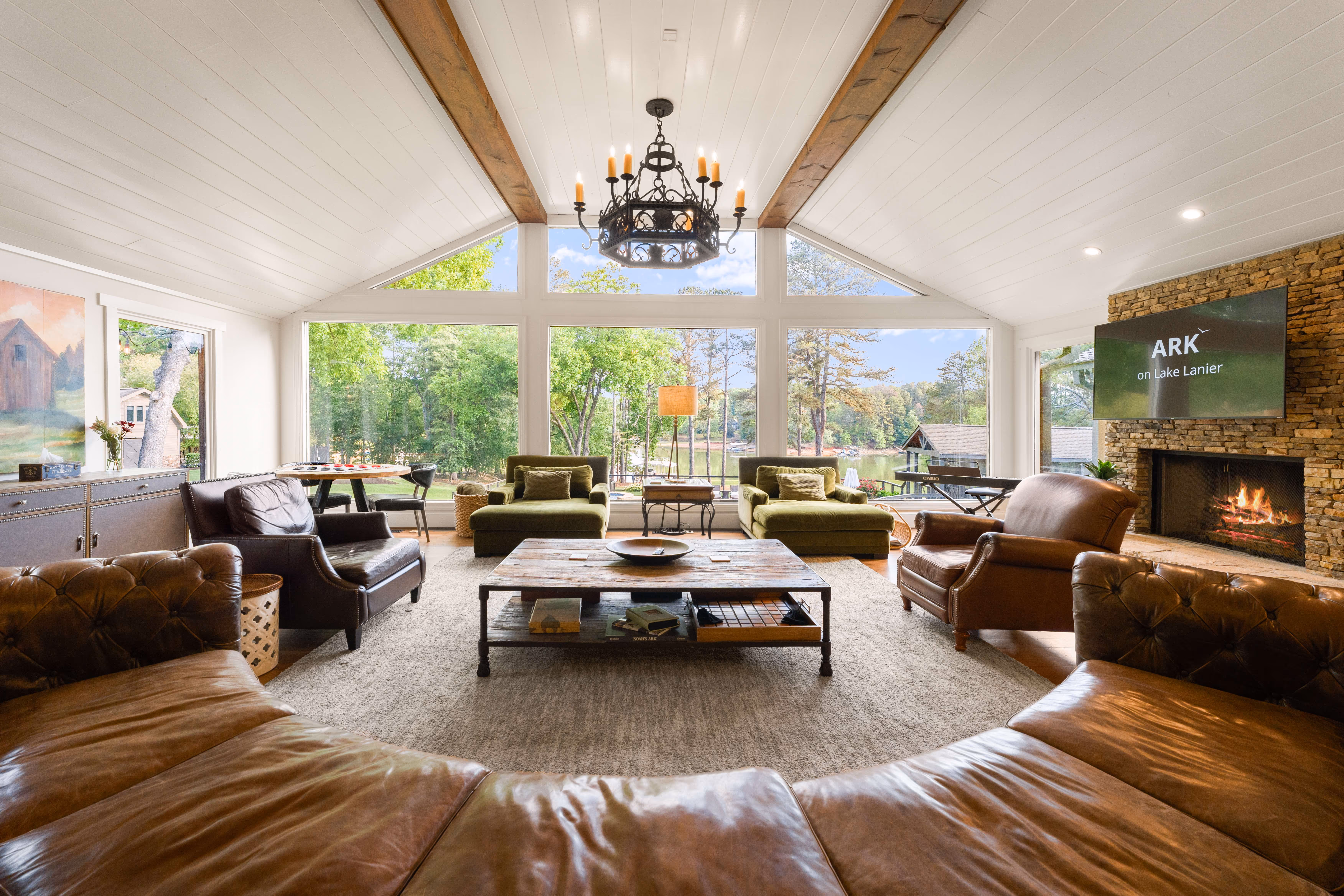 Spacious living room with large windows overlooking trees and a lake, furnished with sofas and armchairs.