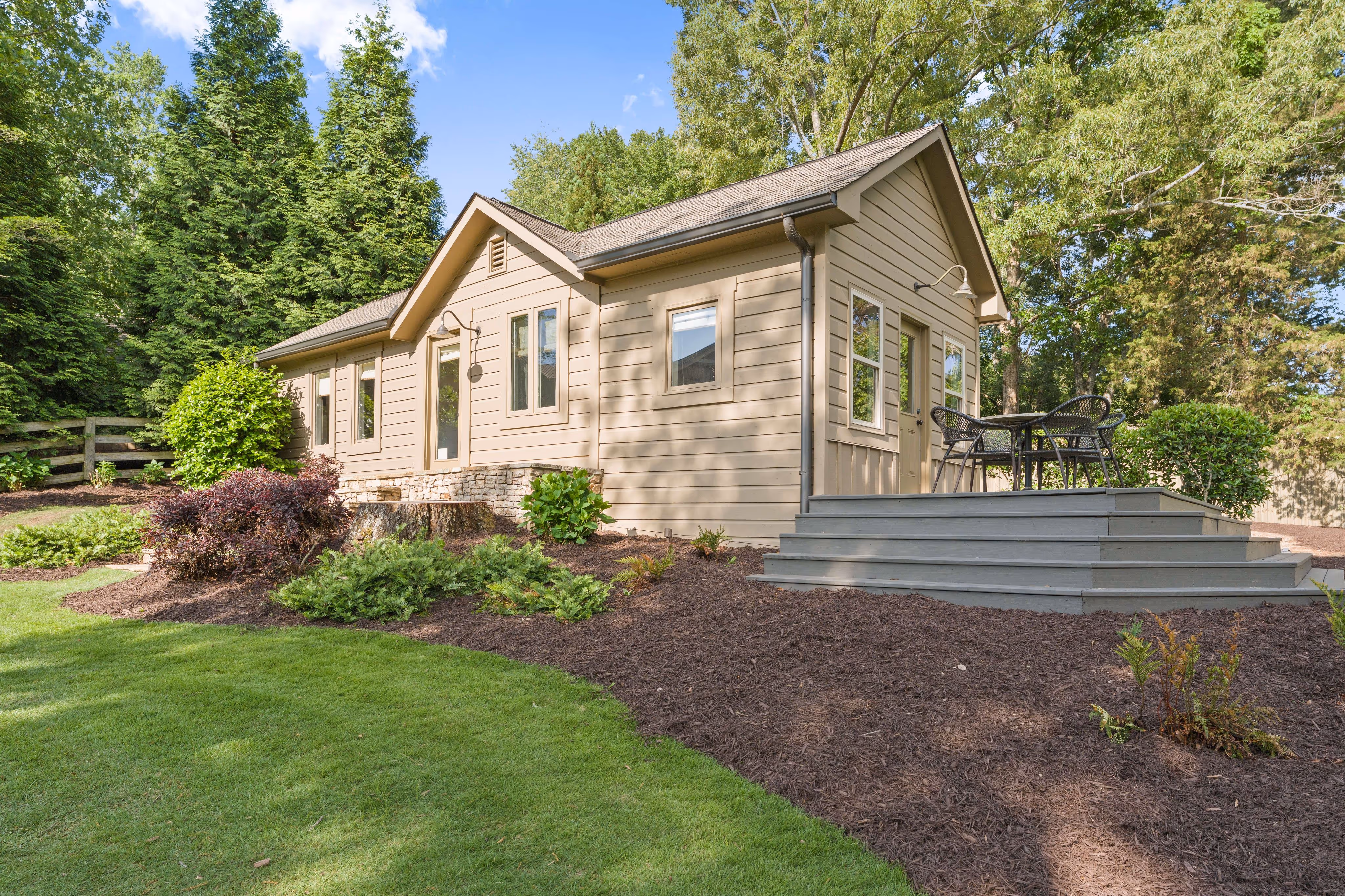 Small beige house with multiple windows, raised deck, outdoor table with four chairs, and surrounding greenery.