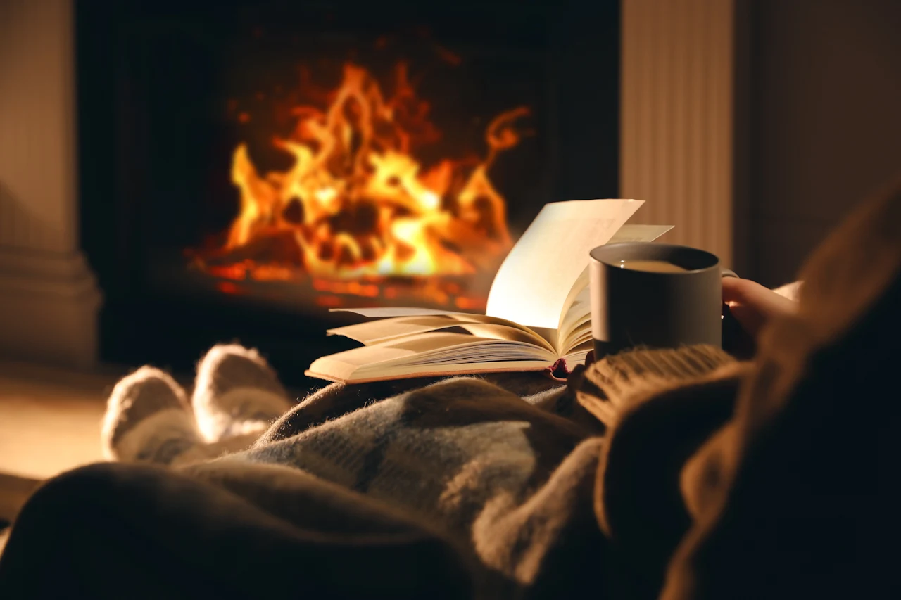 Someone reading a book, and having a coffee while sitting near a fireplace.