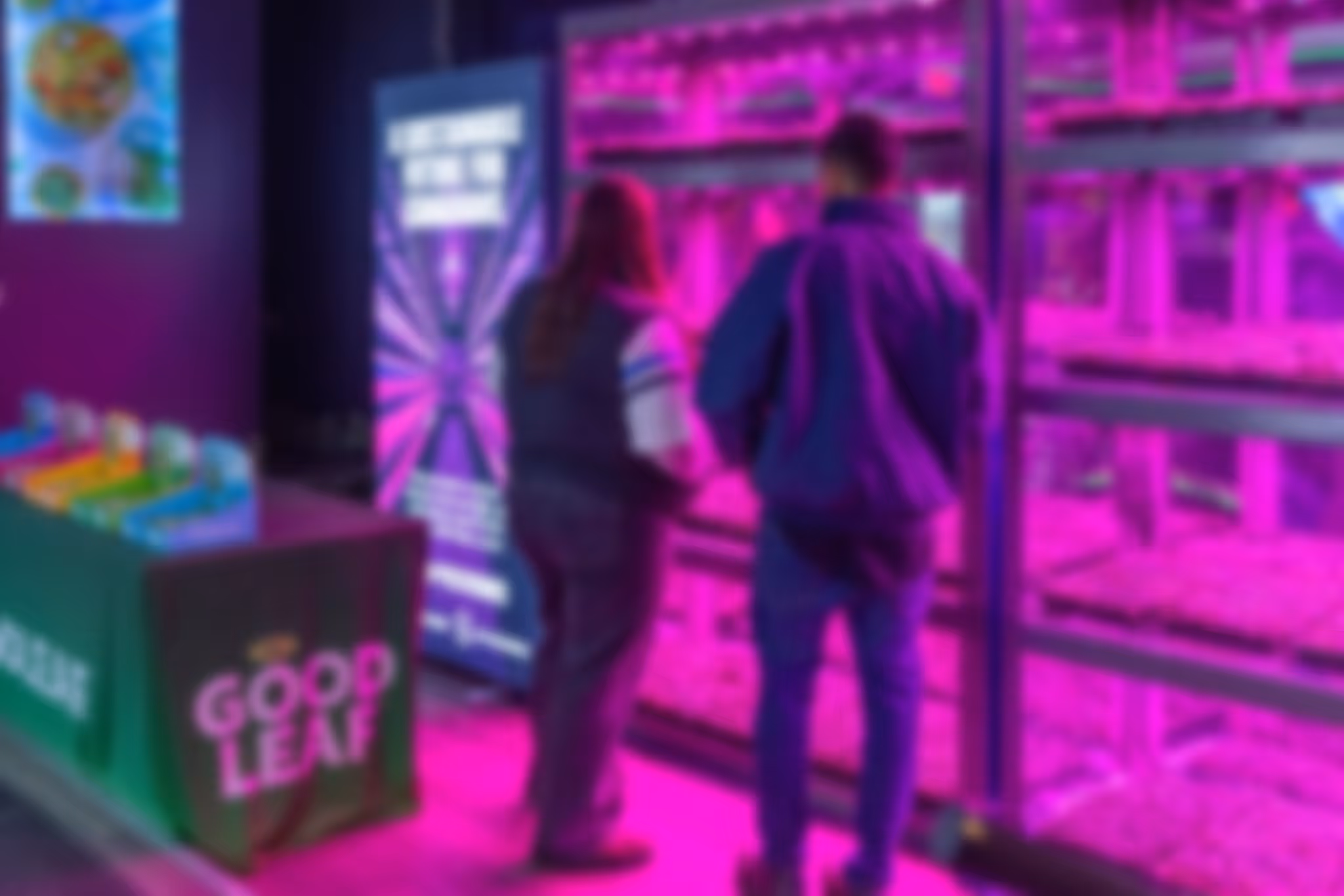 Two people standing in front of indoor vertical farm shelves lit with purple grow lights, next to a table with a green Good Leaf banner.