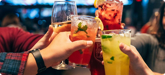 Group of people raising colorful cocktails and drinks in a toast at a lively restaurant.