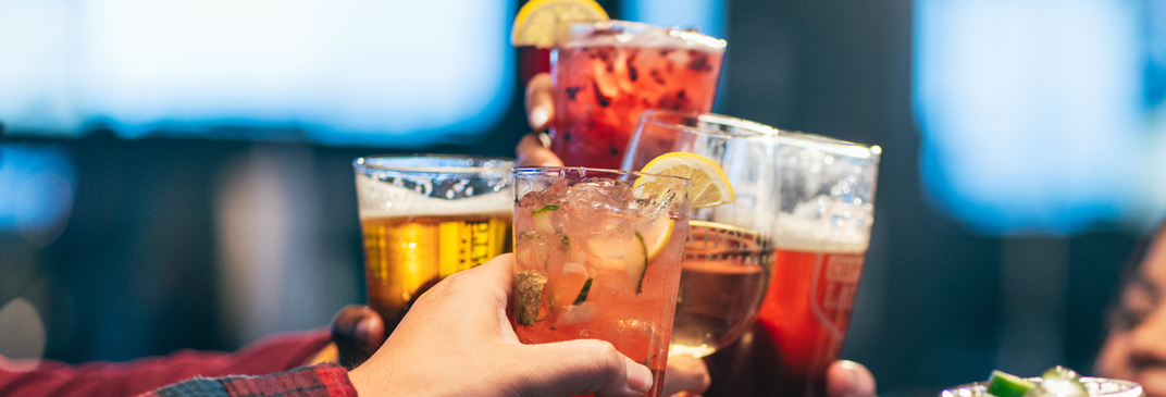 Close up of glasses from group of people raising colorful cocktails and drinks in a toast at a lively restaurant.