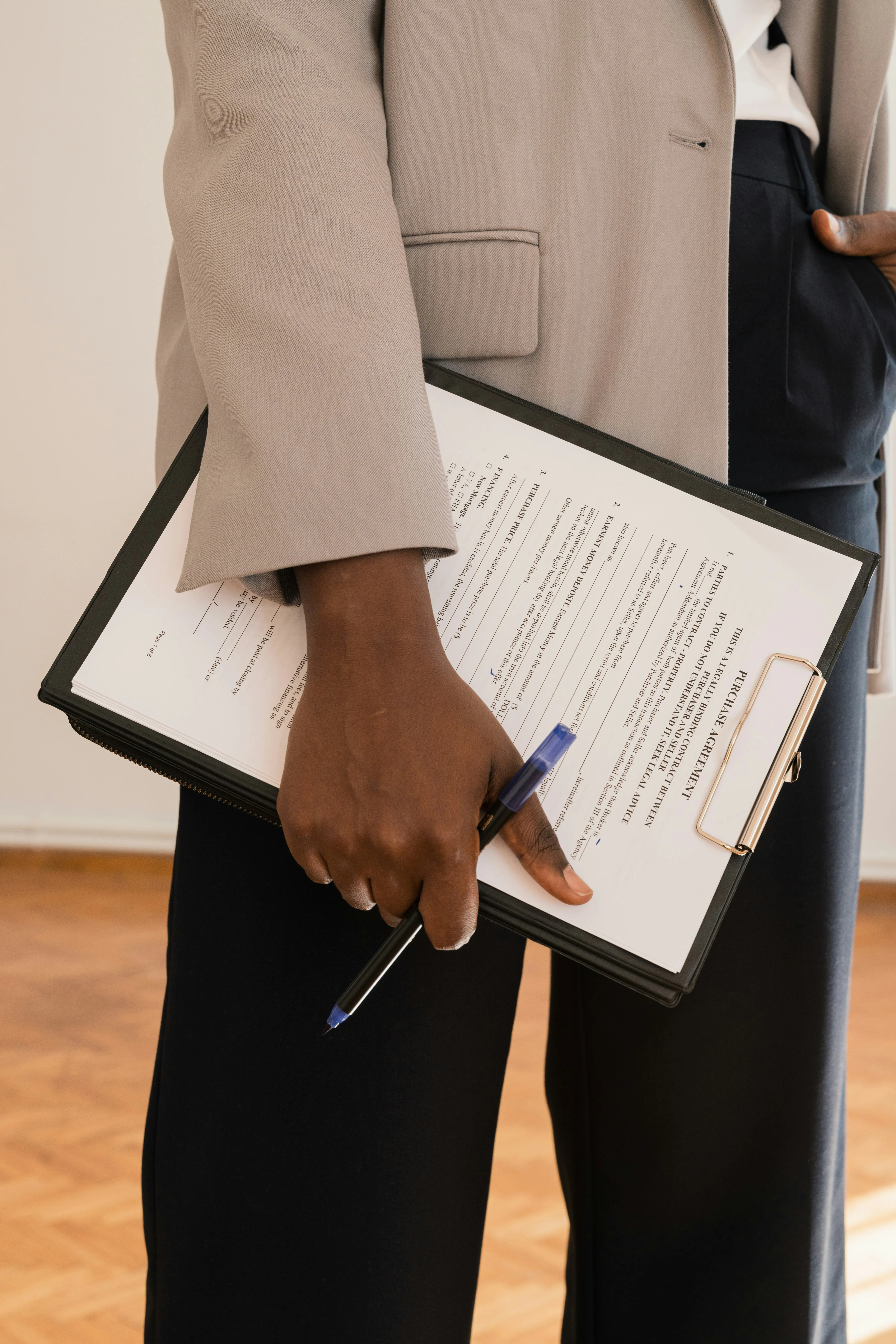 Person holding a clipboard with a purchase agreement document and a blue pen, dressed in a beige blazer and dark pants.