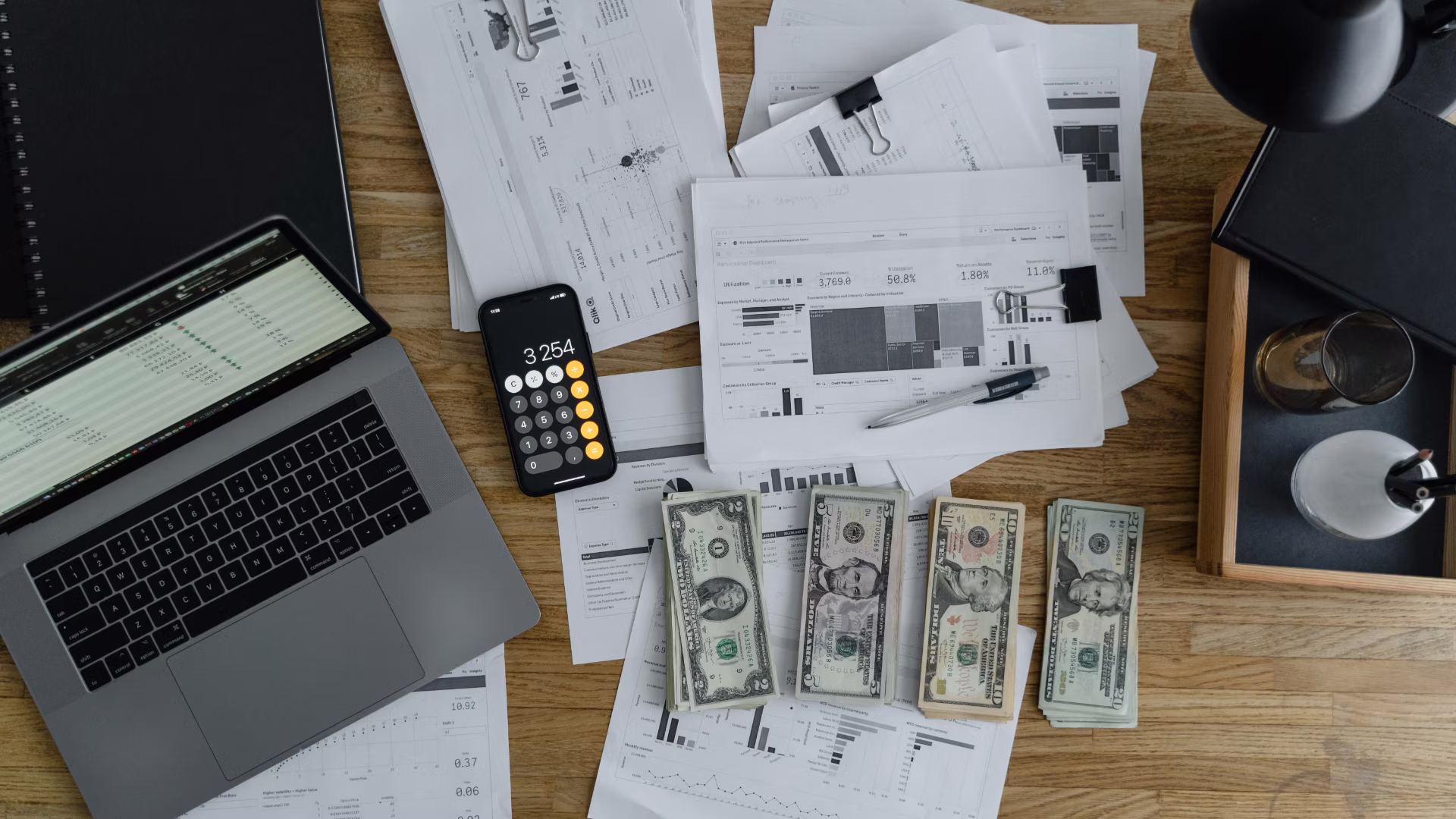 an overhead view of a desk with a laptop, cash, calculator, and pieces of paper scattered across the table