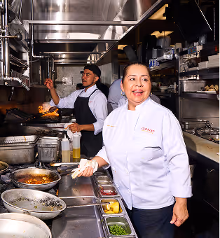 Gloria smiles in the kitchen as fresh-made dishes are being prepared behind her