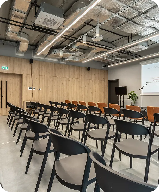 Nowoczesna sala konferencyjna z rzędem czarnych krzeseł, drewnianą ścianą i projektorem. W tle widoczna roślina oraz sprzęt do prezentacji.