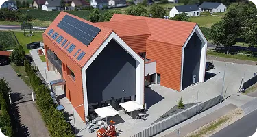 Widok z lotu ptaka na nowoczesny budynek z czerwoną dachówką i fotowoltaiką, otoczony zielonym krajobrazem i parkingiem.