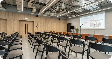 Nowoczesna sala konferencyjna z rzędem czarnych krzeseł, drewnianą ścianą i projektorem. W tle widoczna roślina oraz sprzęt do prezentacji.