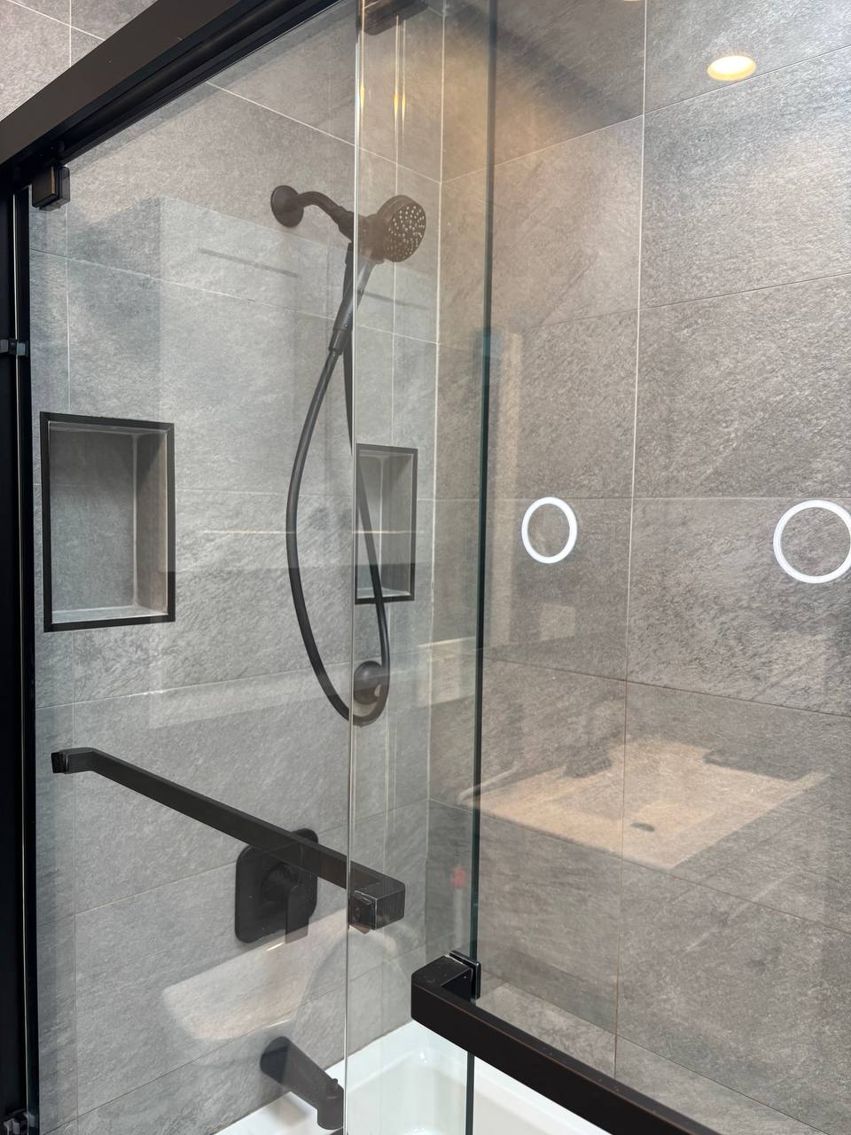 Gray tile shower with black fixtures, hand shower and glass door