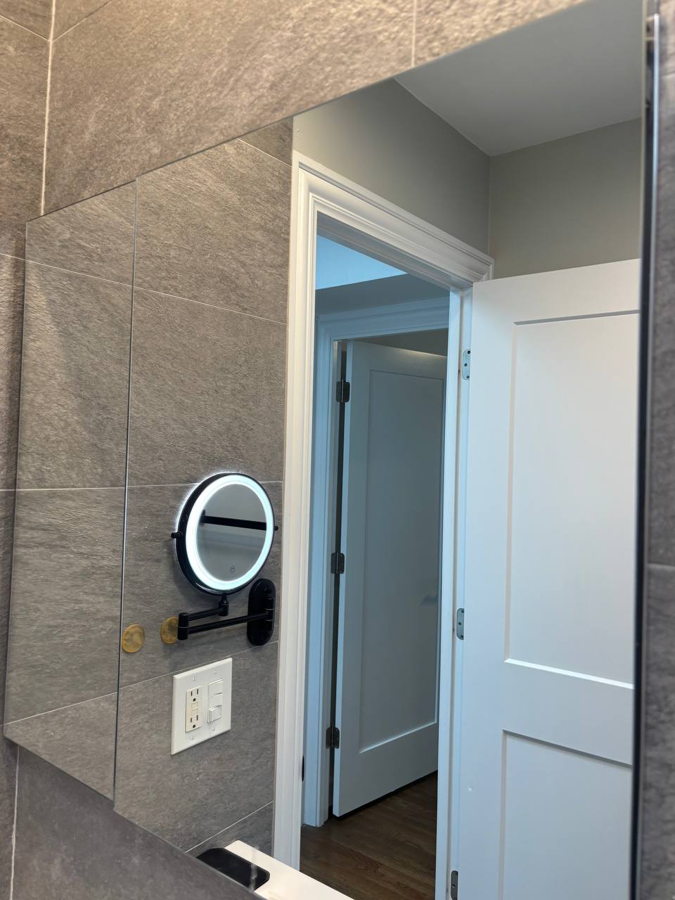 Gray tile bathroom mirror reflecting a lit makeup mirror and doorway