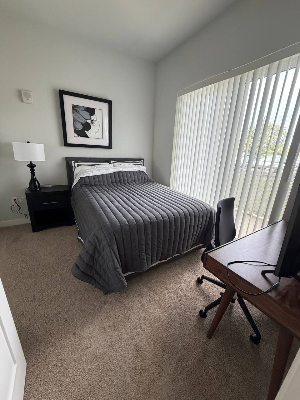 Bedroom with gray quilt, nightstand lamp, desk chair and vertical blinds