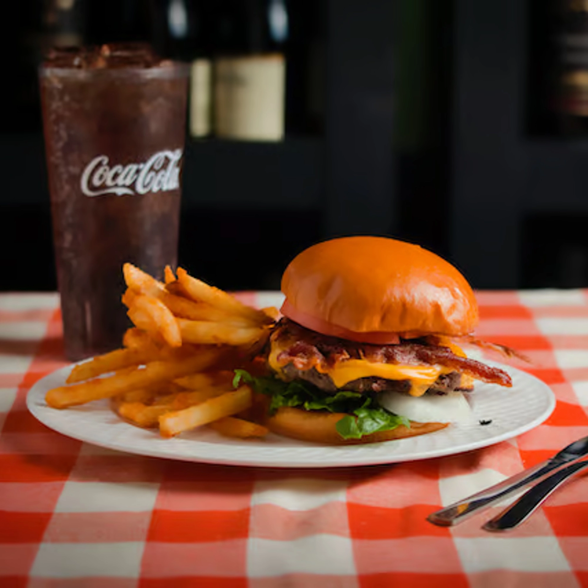 A juicy cheeseburger with lettuce, bacon, and onion sits on a plate with fries and a tall glass of Coca-Cola on a red-checkered tablecloth.