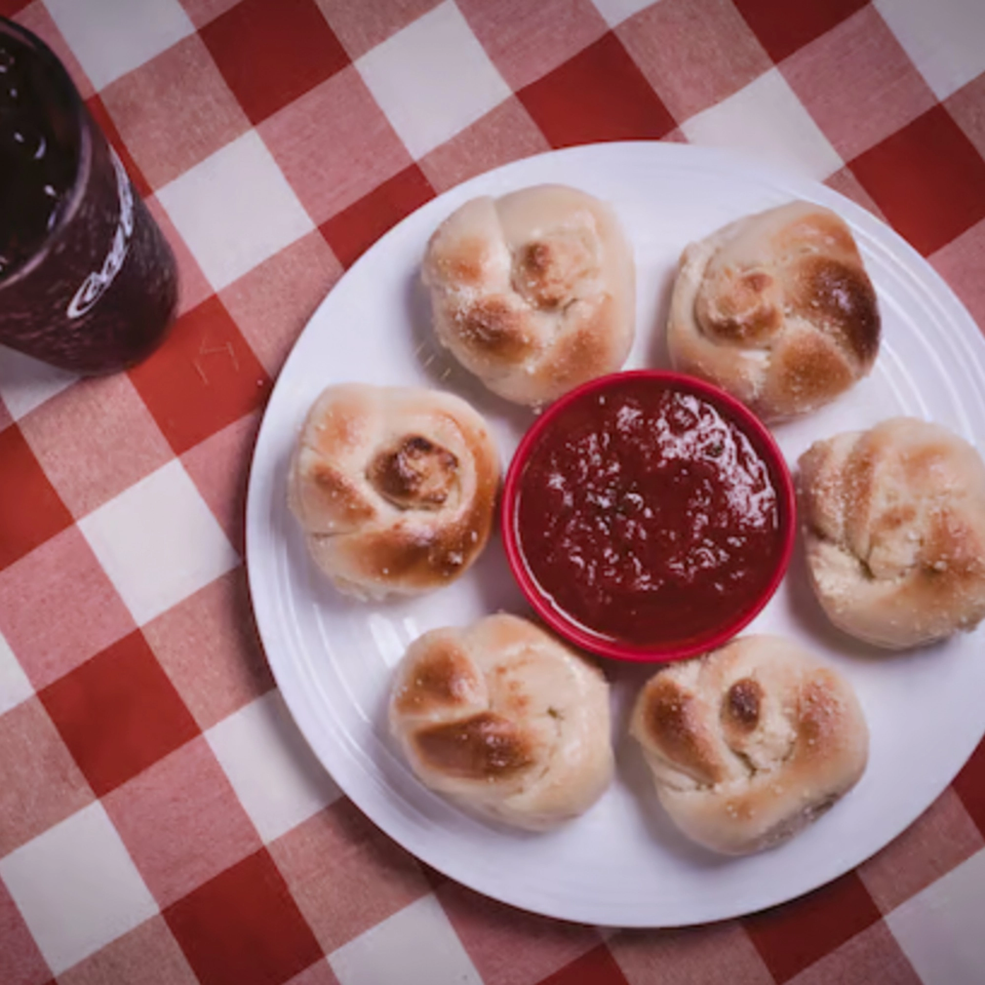 A plate with six garlic knots surrounds a red dish of marinara sauce on a red and white checkered tablecloth. A glass of Coca-Cola is partially visible on the left.