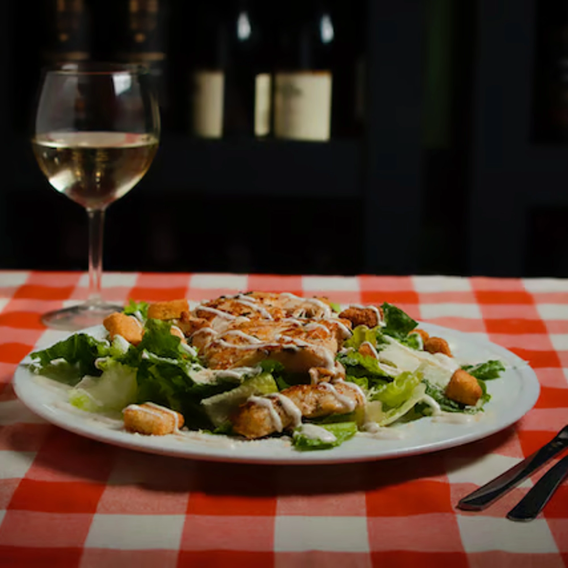 A Chicken Caesar Salad with croutons and dressing is on a white plate, set on a red-checkered tablecloth. A glass of white wine complements the cozy, inviting setting.