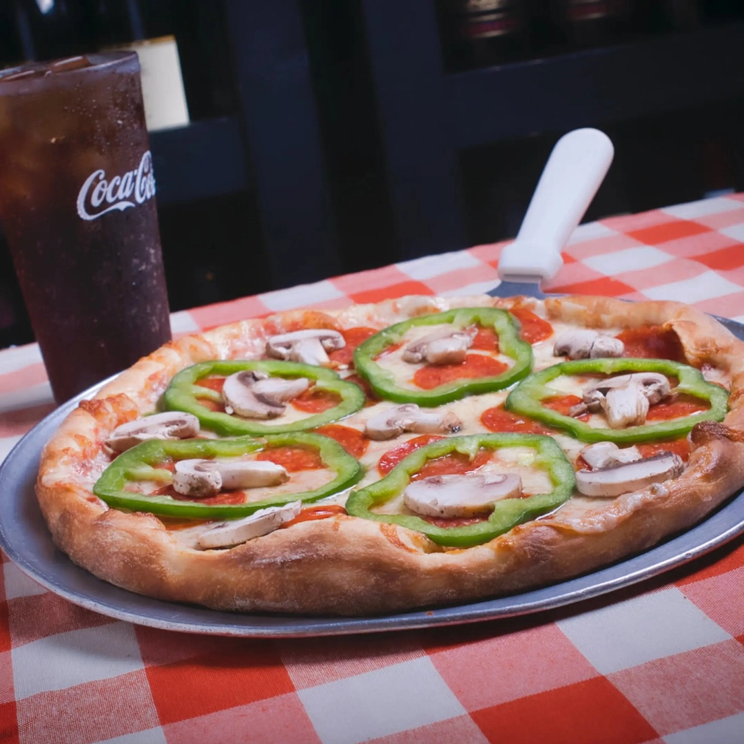 Pizza topped with green pepper rings, mushrooms, and pepperoni on a tray, set on a red-and-white checkered tablecloth. A glass of Coca-Cola is nearby.