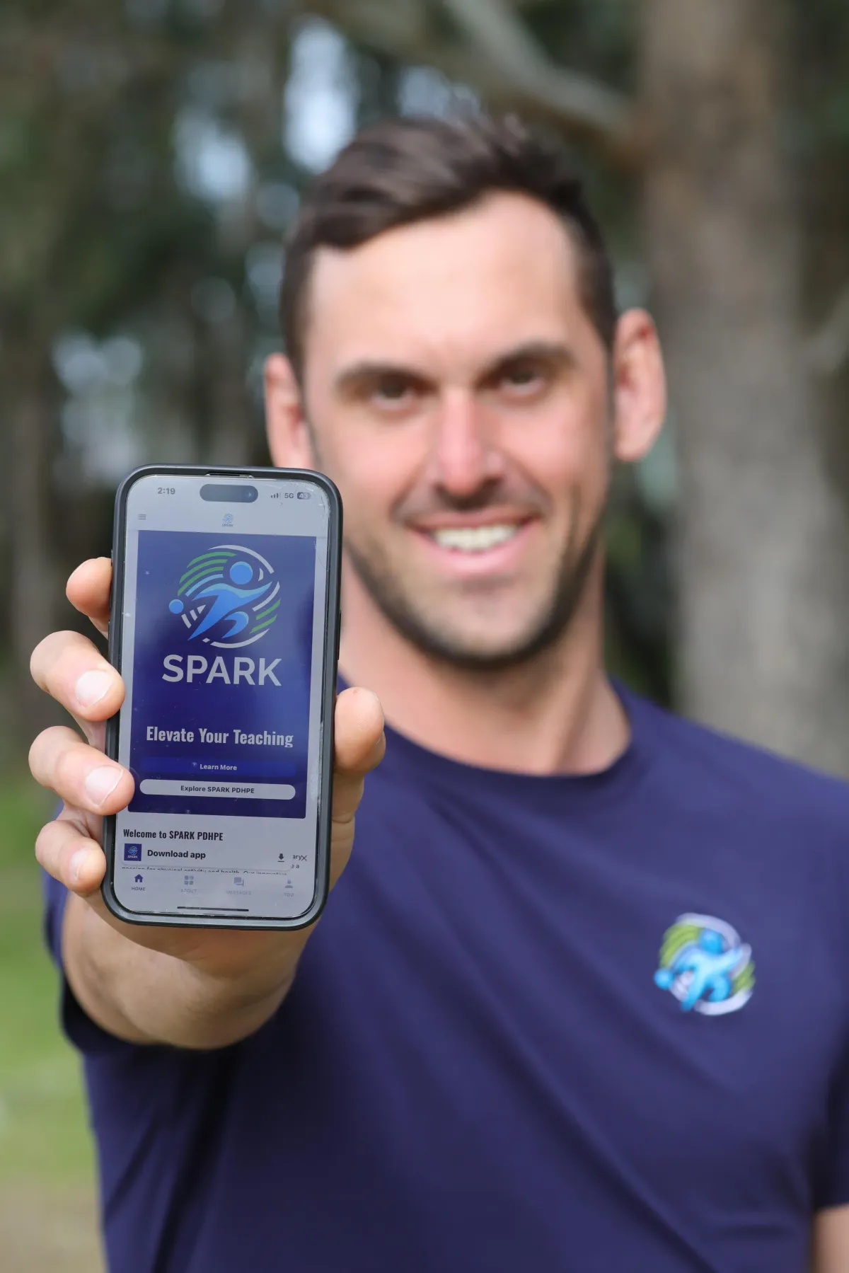A man holding up a phone showing the SPARK PDHPE app
