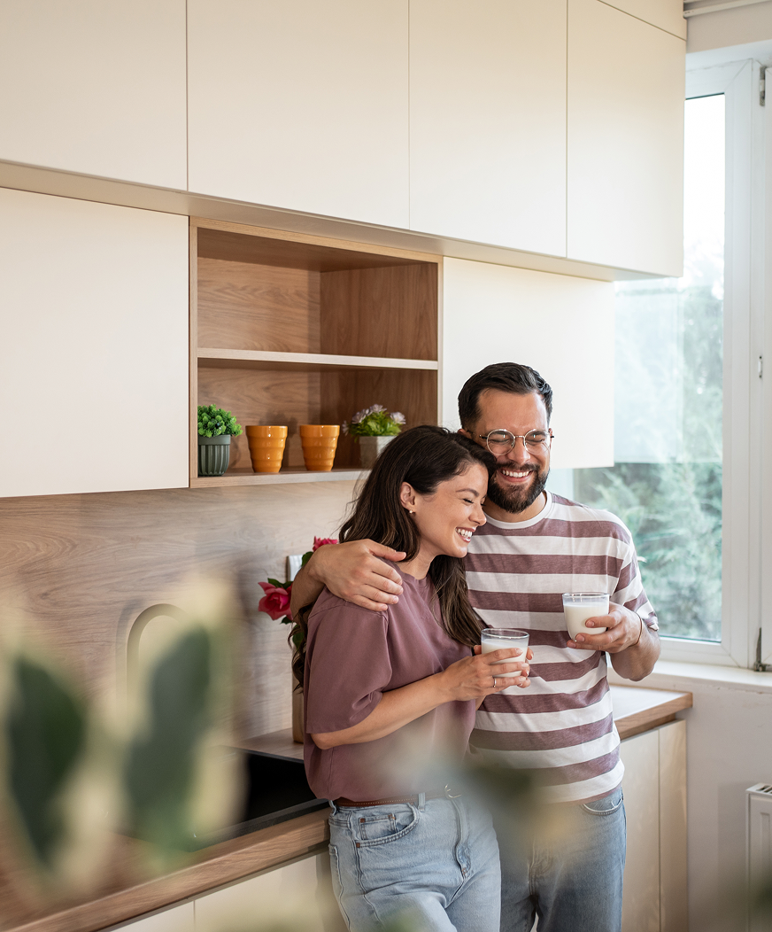A couple stands in a sunny kitchen, holding each other and drinking coffee.
