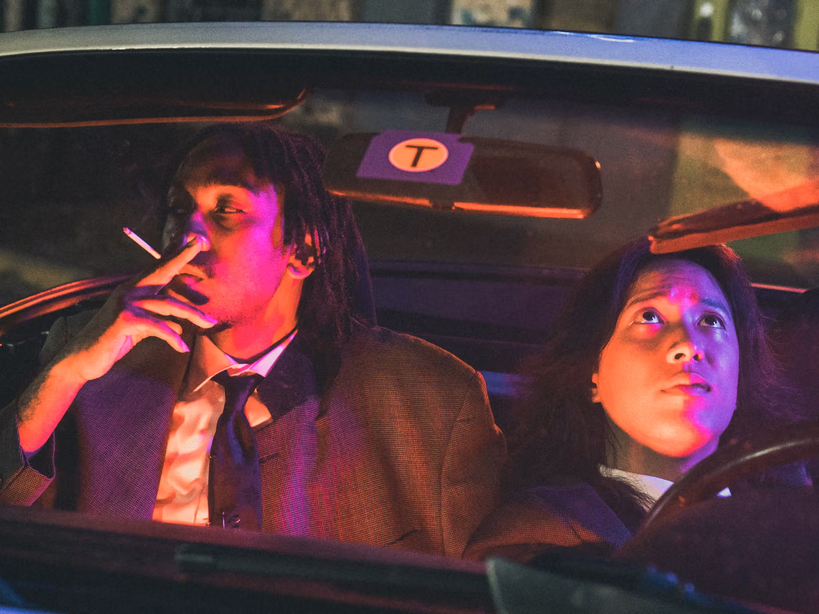 Editorial fashion shot, two models under red neon against a car, one holding a purple lace fan