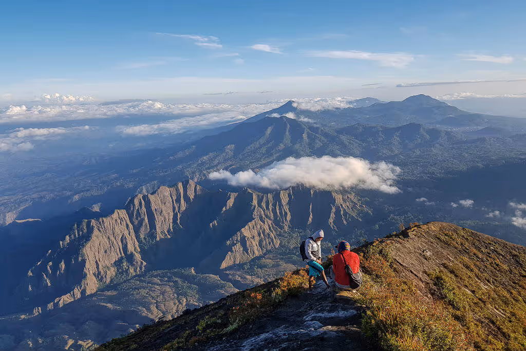 Two hikers sitting on a mountain ridge overlooking a vast landscape of rugged mountains and clouds under a clear blue sky.