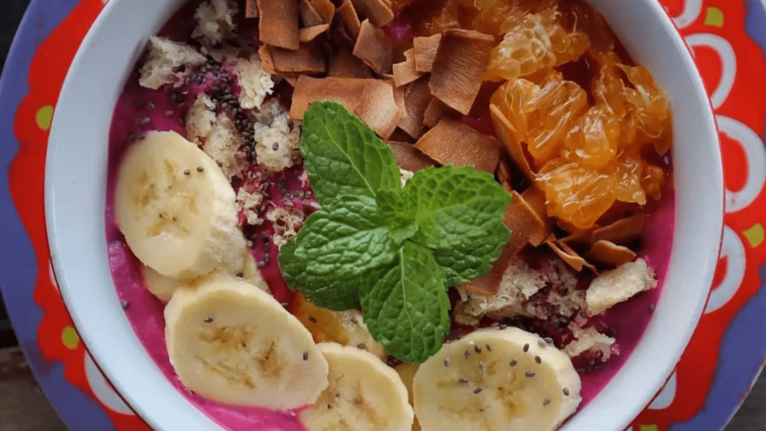 Bowl of pink smoothie topped with banana slices, granola, chia seeds, toasted coconut flakes, orange segments, and a sprig of fresh mint.