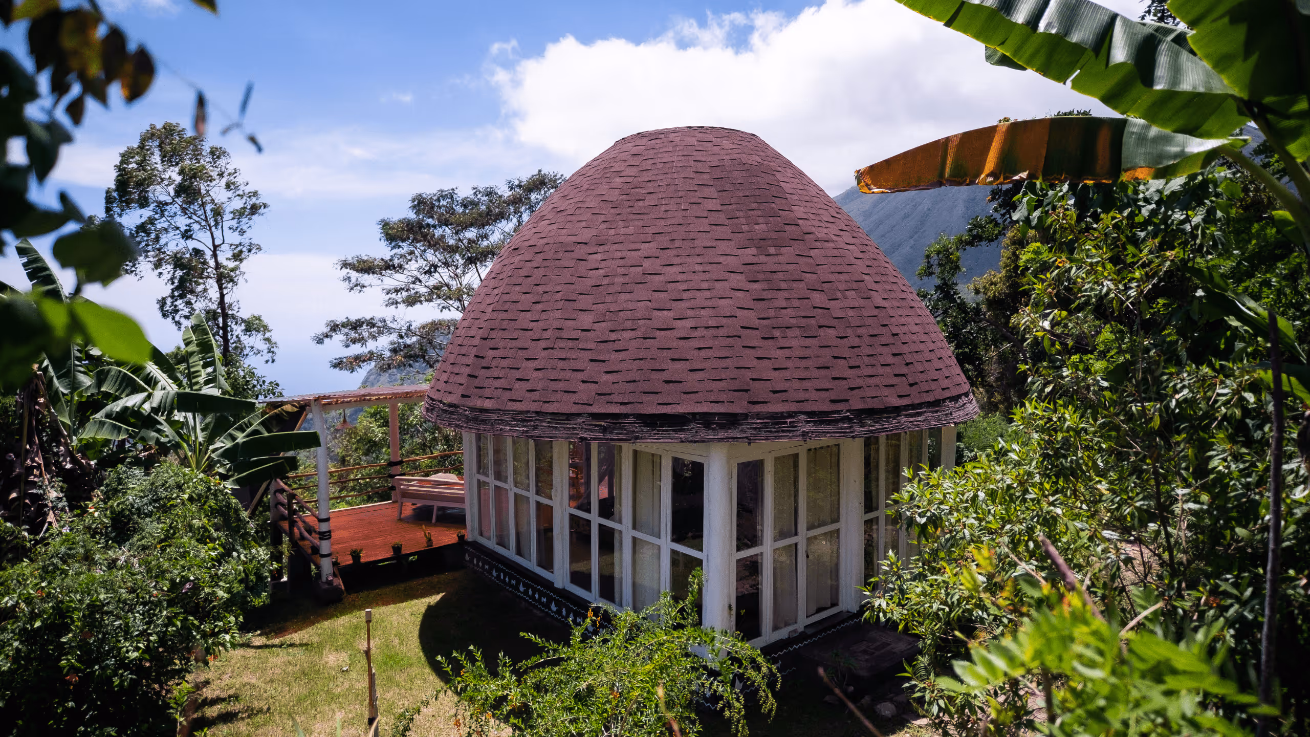 Round cabin with a brown shingle dome roof surrounded by lush green tropical plants and trees.