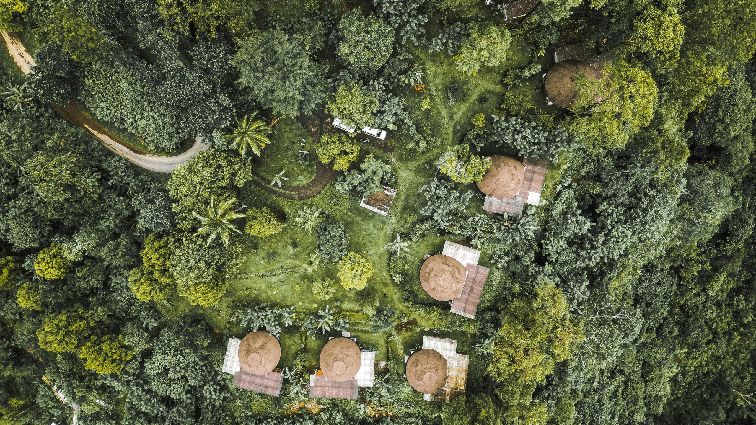 Aerial view of circular thatched-roof huts surrounded by dense green forest and grassy clearings with a narrow dirt path and parked white vehicle.