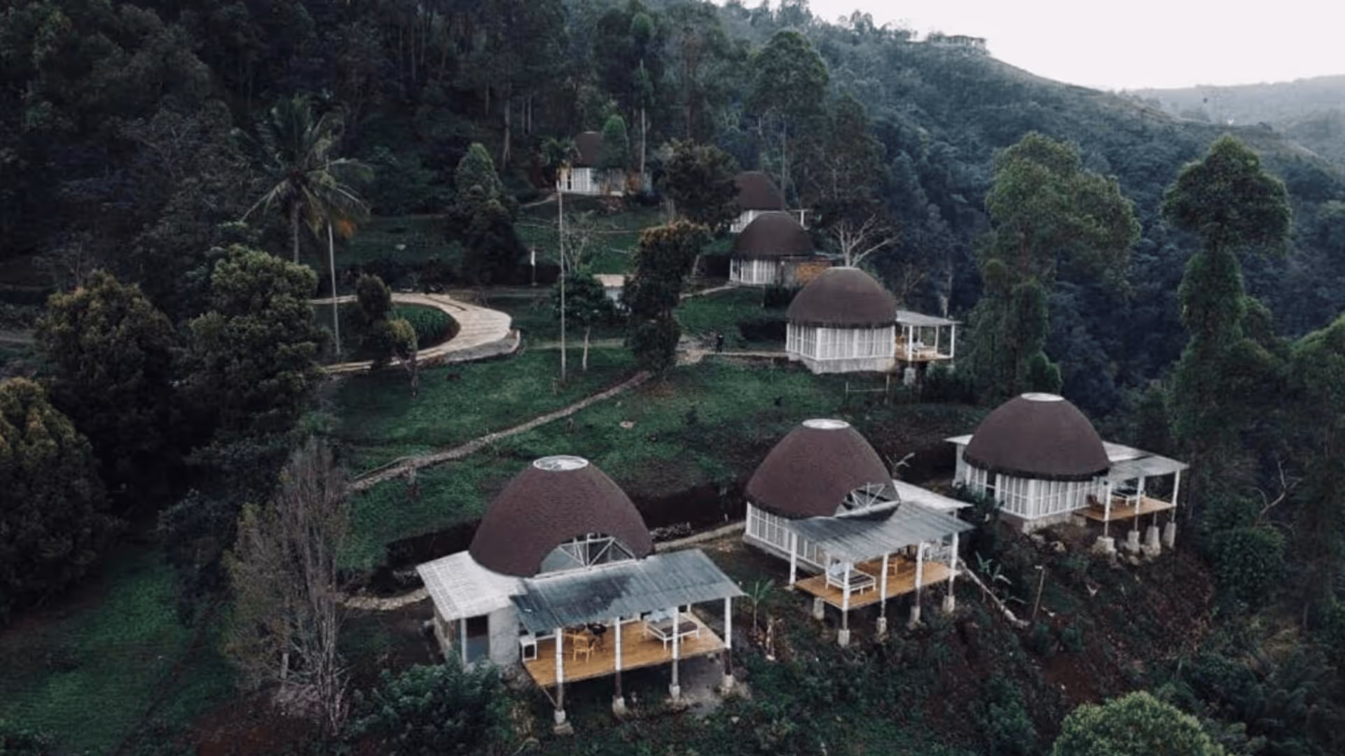 Cluster of dome-shaped cabins with brown roofs and white walls set on a green hillside surrounded by dense trees.